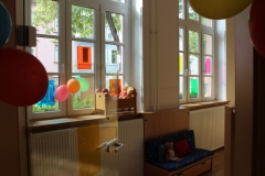 Große Fenster mit Buntpapier und Ballons geschmückt und eine Sitzbank mit Puppenspielzeug.