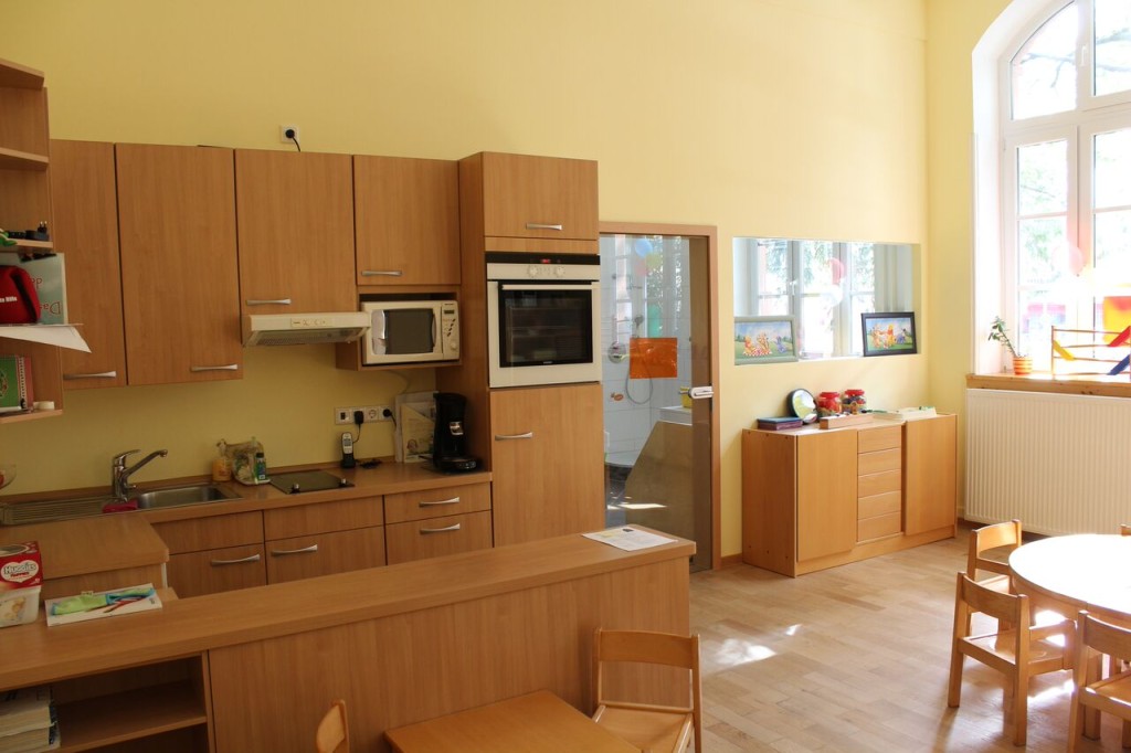Ein heller, gelb gestrichener Raum mit gro´ßßen Fenstern und einer Kindgerchten EInbauküche und anderen Möbelstücken aus Holz.