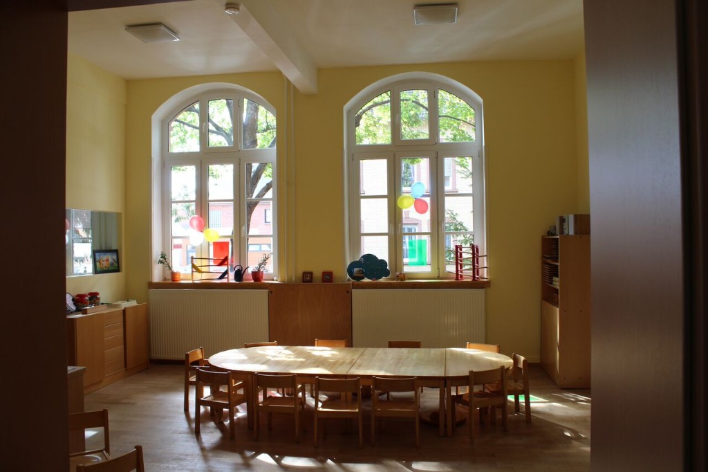Ein heller Raum mit gelben Wänden und großen Fenstern in dem ein Kinderesstisch mit Platz für 12 Kinder steht.